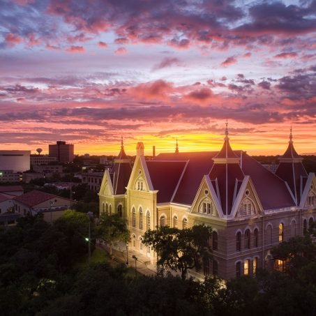 Old Main at Texas State University glowing at sunset with a colorful pink and orange sky over the San Marcos campus.